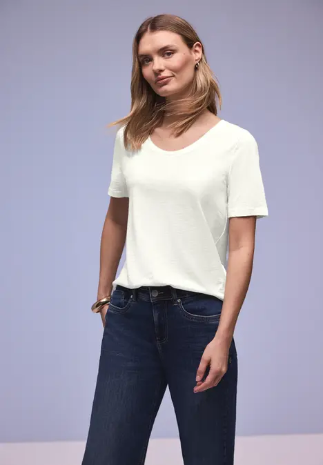 Eine Frau trägt ein schlichtes weißes T-Shirt und dunkle Jeans vor neutralem Hintergrund.