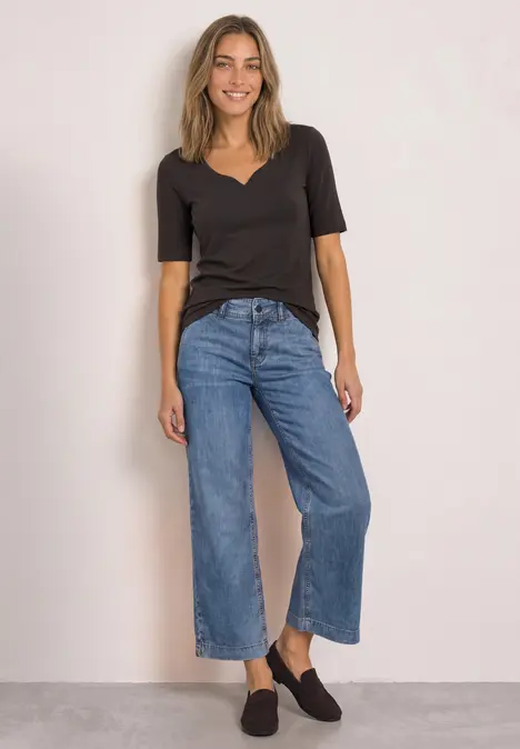 Frau trägt schwarzes V-Neck-Shirt und weite, taillierte Culotte-Jeans vor hellem Hintergrund.