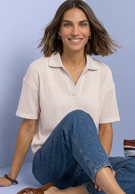 Eine lächelnde Frau trägt Poloshirt und Jeans, sitzt entspannt mit verschränkten Beinen.