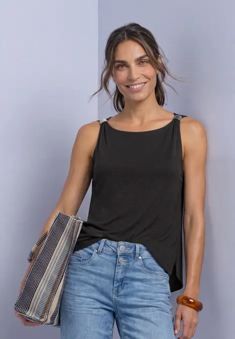 Lächelnde Frau in schwarzem Top und Jeans hält gefaltetes Strickjäckchen.