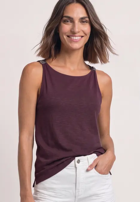 Frau lächelt in weinrotem ärmellosem Top und weißer Hose, Hand in der Tasche.