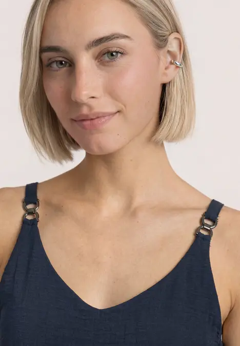 Junge Frau mit kurzem blondem Bob trägt dunkelblauen Top mit Riemen und Ohrschmuck vor hellem Hintergrund.