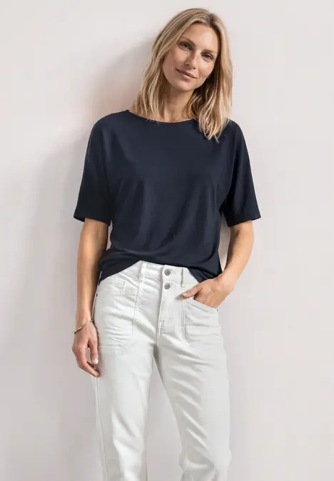 Frau in dunkelblauem T-Shirt und weißen Jeans, steht seitlich mit einer Hand in der Hosentasche.
