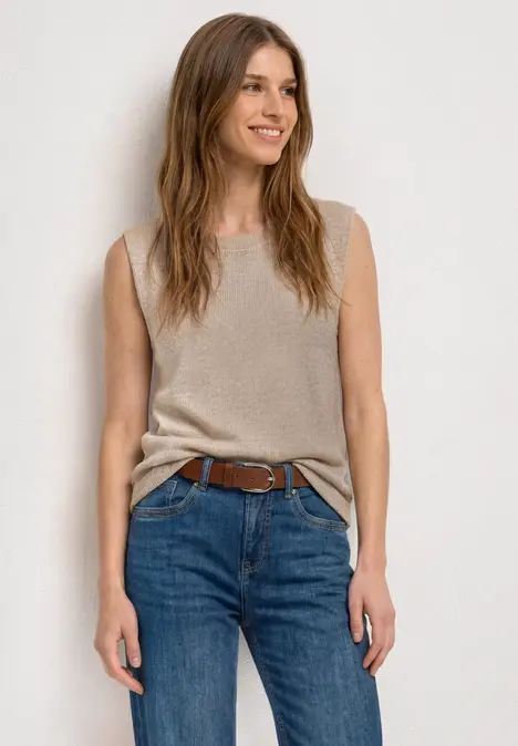 Frau in ärmellosem beigem Top mit Knoten und blauen Jeans vor hellem Hintergrund.