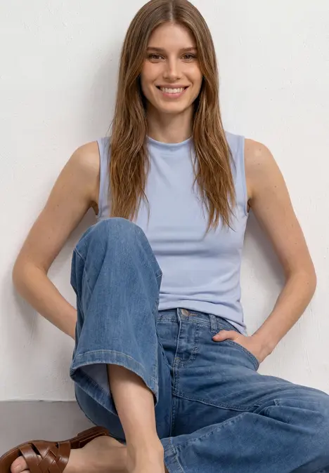 Eine Person sitzt vor weißem Hintergrund, trägt ein hellblaues Top, blaue Jeans und braune Sandalen.