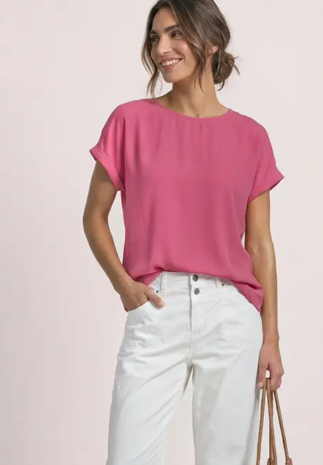 Frau trägt pinkes kurzärmeliges Oberteil und weiße Hose, lächelt, Hand in Tasche, lässiger Look.