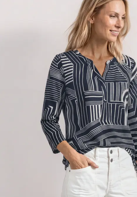 Frau trägt eine dunkelblaue Bluse mit weißem, geometrischem Muster und 3/4-Ärmeln, kombiniert mit weißen Jeans.