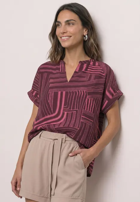 Frau in pink-violett gemustertem Oberteil und beige Paperbag-Hose vor hellem Hintergrund.