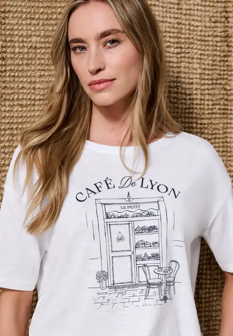 Person trägt weißes T-Shirt mit Aufdruck 'Café de Lyon' und Café-Skizze vor geflochtenem Hintergrund