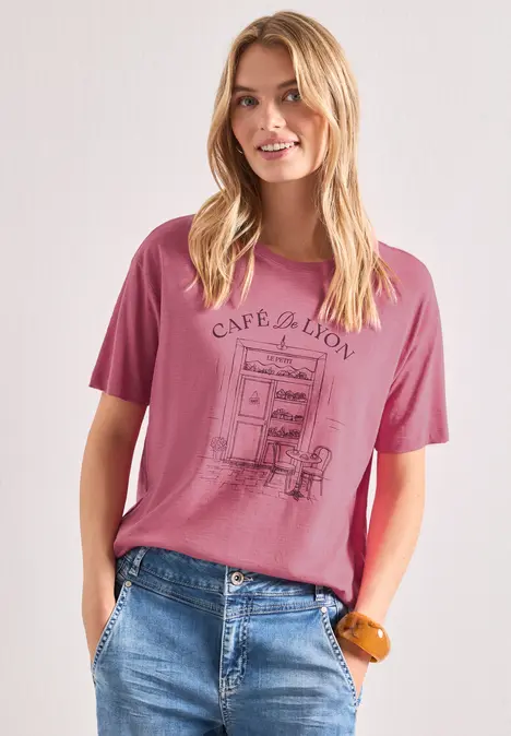 Blonde Frau trägt pinkes T-Shirt mit Café-Motiv und blaue Jeans, mit Händen in den Taschen.
