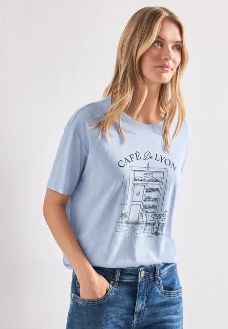 Frau in hellblauem T-Shirt mit 'CAPE DE LYON'-Aufdruck und Jeans vor weißer Wand