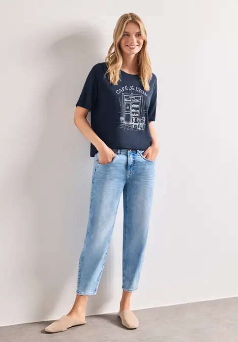 Frau in navy Shirt mit Print, hellblauen Jeans und beigen Slippern steht vor weißer Wand, Hände in den Taschen.