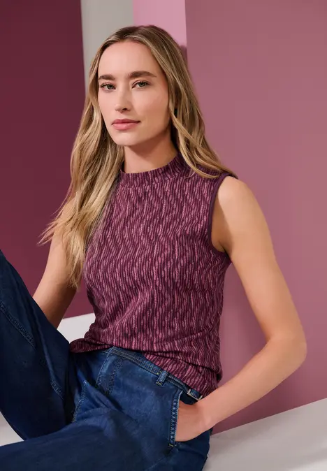 Frau mit locker sitzender lila Strickweste und Jeans lehnt vor rosa Hintergrund.