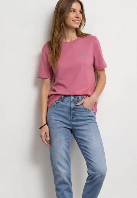 Frau in rosa T-Shirt und blauen Jeans steht lächelnd vor weißem Hintergrund.