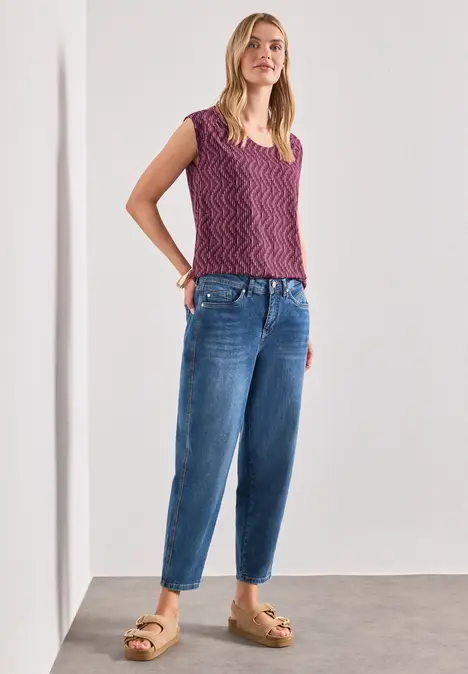 Frau in lockeren Jeans, lila Stricktop und Plateausandalen steht vor weißer Wand.