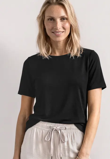 Frau in schwarzem T-Shirt und beiger Hose mit Kordelzug vor neutralem Hintergrund.