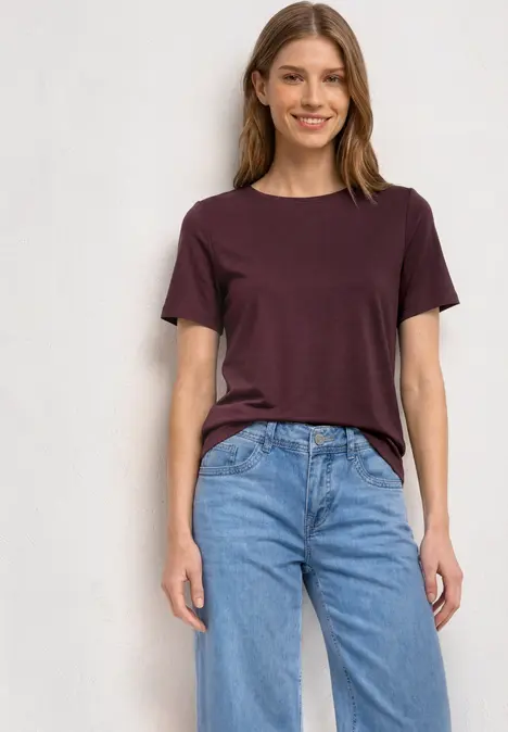 Eine junge Frau in lila T-Shirt und blauen Jeans steht lächelnd vor neutralem Hintergrund.