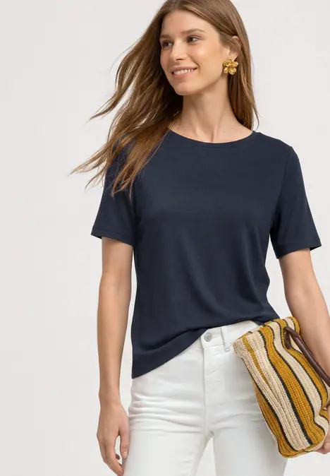 Frau trägt dunkelblaues T-Shirt, weiße Hose und hält gestreifte Clutch, lächelt vor hellem Hintergrund