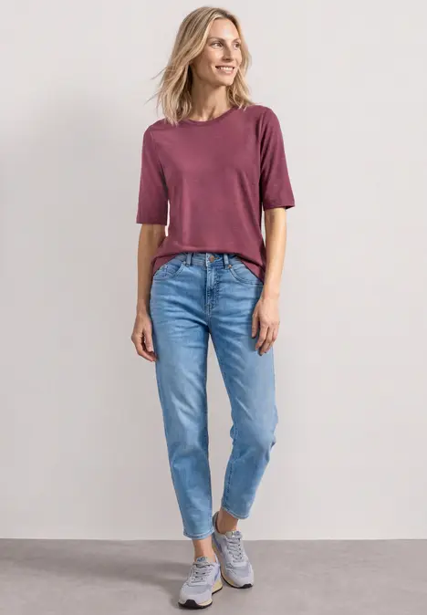 Eine junge Frau trägt ein rotes T-Shirt, blaue Jeans und graue Sneaker vor schlichter Wand.