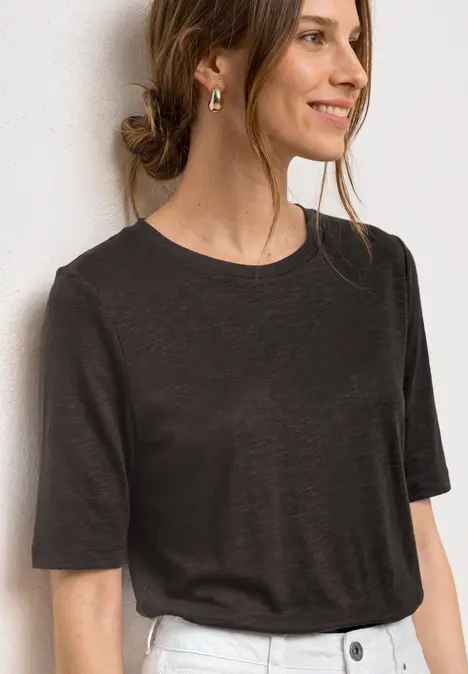 Frau in dunkelgrauem T-Shirt mit goldenem Ohrring und Halskette vor weißem Hintergrund
