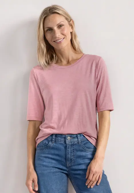 Frau im rosa T-Shirt und blauen Jeans vor hellem Hintergrund.