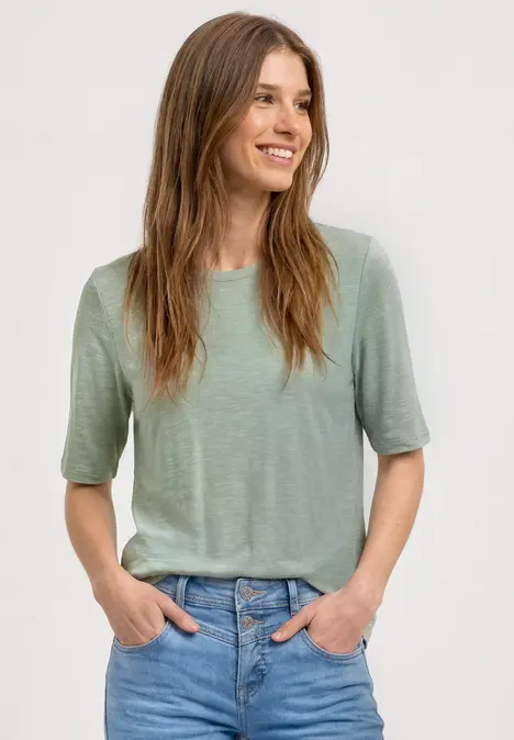 Frau mit unkenntlich gemachtem Gesicht trägt grünes T-Shirt und Jeans, Hände in den Taschen