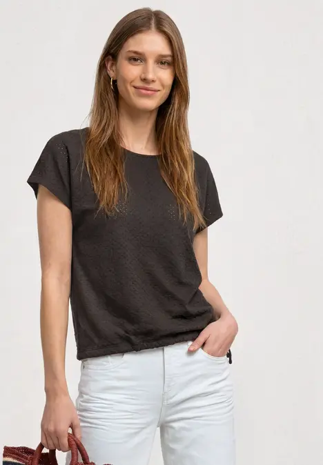 Junge Frau im dunklen T-Shirt und weißen Jeans, lächelnd vor hellem Hintergrund.