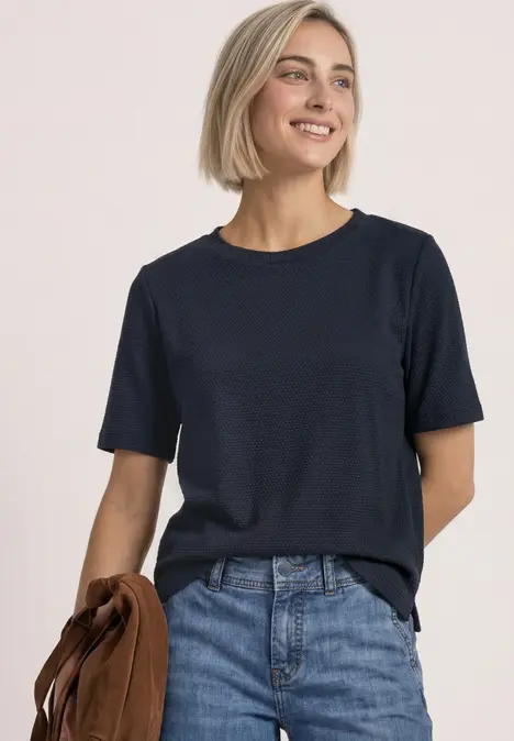 Frau mit kurzem blondem Haar in dunkelblauem T-Shirt und Jeans, hält braune Jacke und lächelt.
