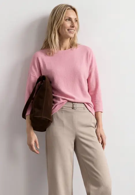 Eine lächelnde Frau in beiger Hose, rosafarbenem Pullover und brauner Tasche.
