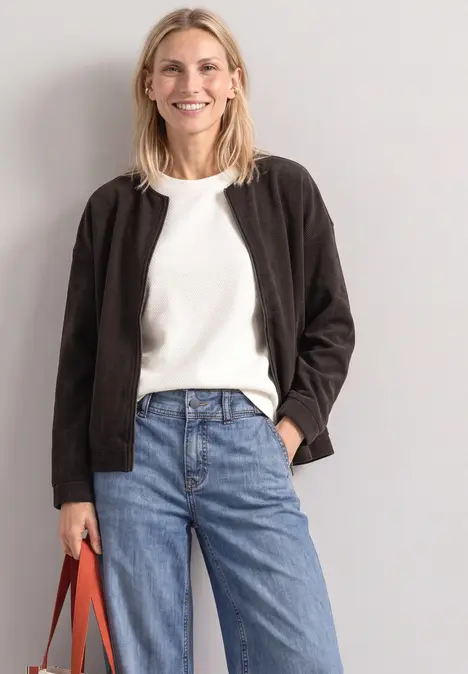 Eine lächelnde Frau trägt schwarzen Blouson, weißen Pullover, blaue Jeans und hält eine rote Tasche.