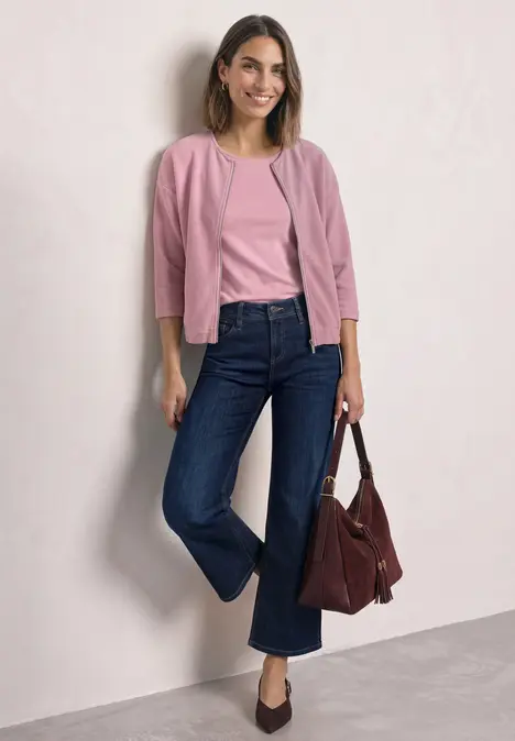 Eine lächelnde Frau lehnt an der Wand, trägt rosa Oberteile, dunkelblaue Jeans und hält eine dunkelrote Tasche.