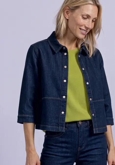 Eine Frau trägt ein dunkles Jeans-Jackett und Jeanshose mit grünem Shirt, lächelt nach unten.