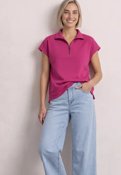 Eine lächelnde Frau trägt ein pinkes Polo-Shirt mit Reißverschluss und helle, weite Jeans.