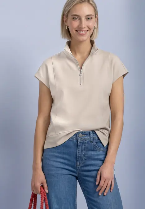 Eine lächelnde Frau in beigeem Zip-up-Top und Jeans hält eine rote Tasche.
