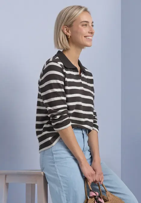 Eine lächelnde Frau mit kurzem blondem Haar, gestreiftem Pullover und hellblauer Jeans lehnt sich seitlich ab.