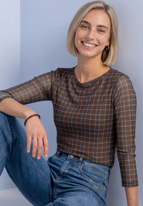 Lächelnde Frau mit kurzen blonden Haaren in kariertem Oberteil und Jeans sitzt vor hellem Hintergrund.