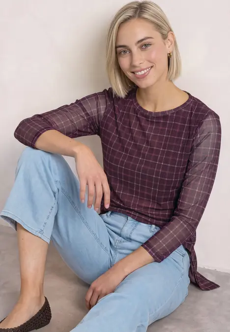 Eine lächelnde Frau in kariertem Pullover und Jeans sitzt entspannt auf dem Boden.