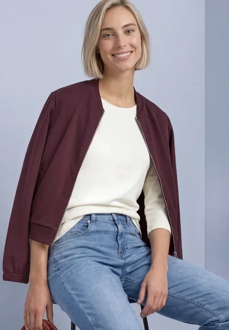 Junge Frau sitzt lächelnd auf einem Hocker, trägt weiße Bluse, Jeans und eine weinrote Jacke locker über den Schultern.
