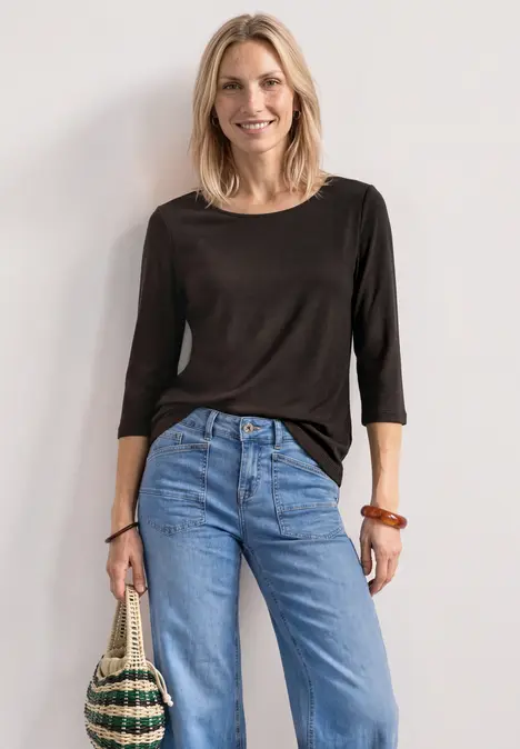 Lächelnde Frau in schwarzem Oberteil und blauen Jeans, hält eine Strohtasche vor hellem Hintergrund