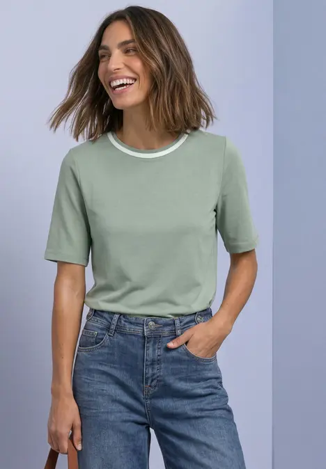 Frau lächelt, trägt grünes T-Shirt mit weißem Kragen und Jeans, steht mit einer Hand in der Tasche.