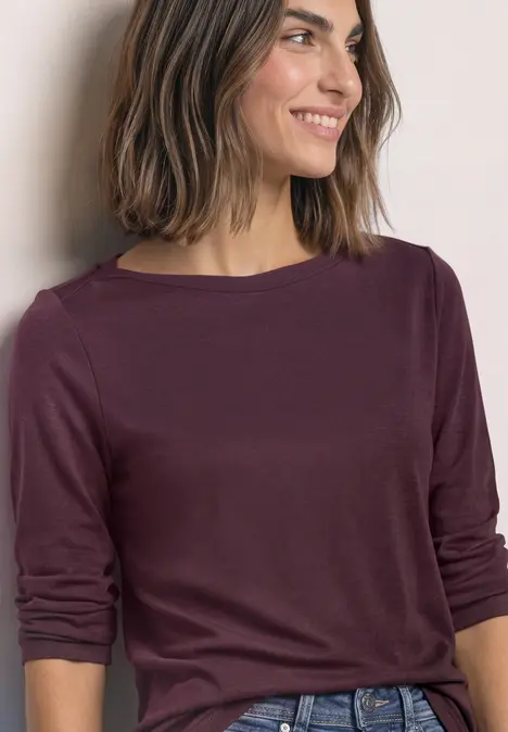 Frau mit schulterlangen Haaren in weinrotem Langarmshirt, lächelt seitlich.