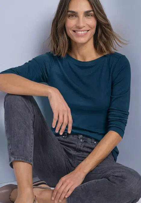 Frau in langärmeligem, dunkelblauem Oberteil und Jeans sitzt lächelnd mit einem angewinkelten Bein.