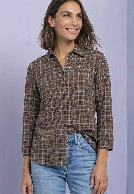 Eine Frau steht lächelnd vor neutralem Hintergrund in einem braunen karierten Hemd und blauen Jeans.