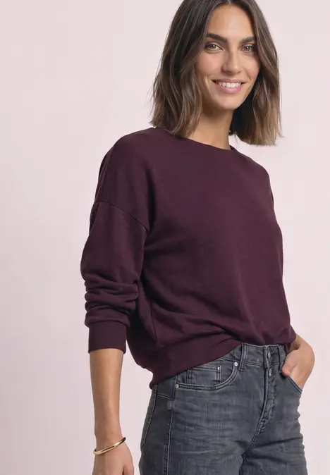 Een glimlachende vrouw draagt een bordeauxrode trui en grijze jeans tegen een lichte achtergrond.