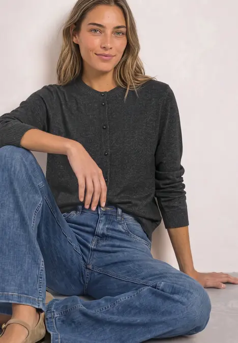 Eine lächelnde Frau in dunkelgrauem Cardigan und blauen Jeans sitzt entspannt vor hellem Hintergrund.