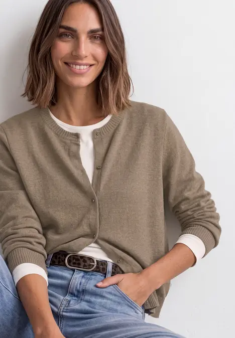 Model lacht zittend in jeans, wit shirt en kaki vest, gecombineerd met een riem.