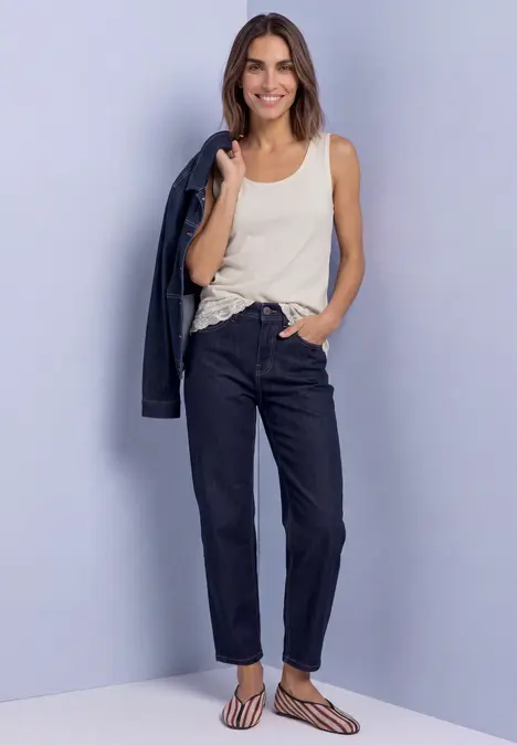 Vrouw in crèmekleurig topje en donkere jeans, glimlachend met jasje over schouder.