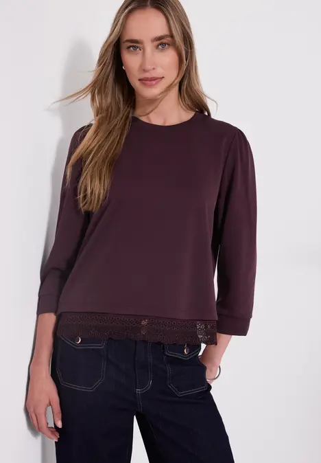 Eine Frau in dunkelviolettem Langarmshirt mit Spitzensaum und blauer Jeans.
