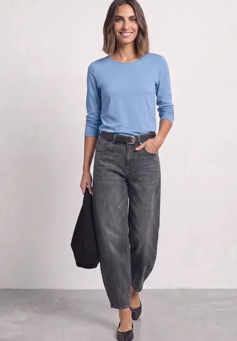 Vrouw in blauw shirt met lange mouwen, losse zwarte spijkerbroek en zwarte schoenen, met tas.