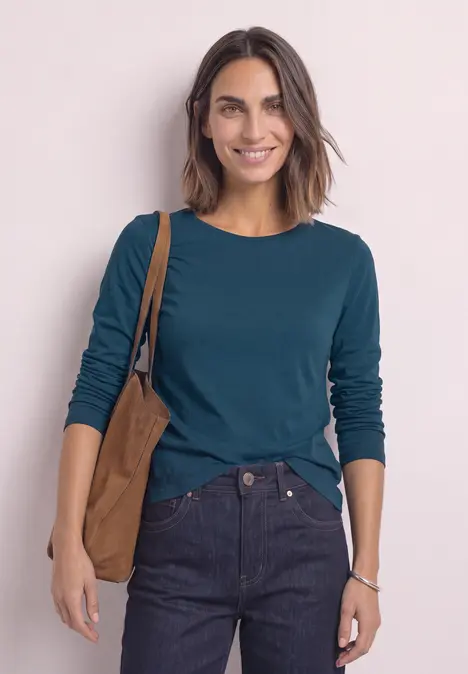 Frau mit dunklen Haaren in dunkelgrünem Langarmshirt und Jeans, lächelnd mit brauner Tasche